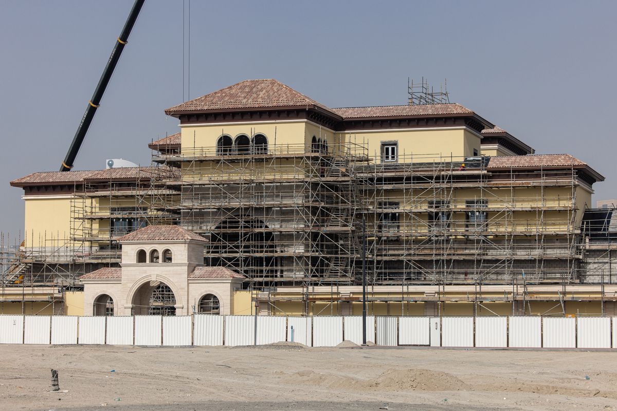 A long stretch of tan sand in front of a white construction fence foregrounds a massive yellow villa with a brown-tiled roof and an arched gateway. All around it is construction scaffolding that reaches up several stories, with a large crane in the background.