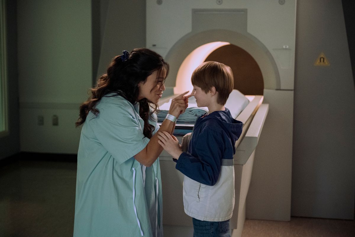 gina rodriguez wearing a hospital gown, leaning to touch a young boy on the nose, while in front of an MRI