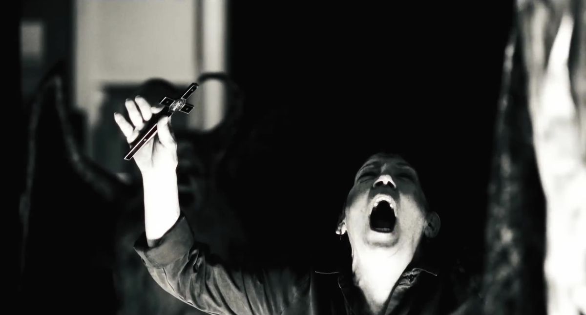 Ann Dowd in the The Exorcist: Believer holding up a cross and shouting in a dark room, with the very vague shadow of a winged figure silhouetted behind her