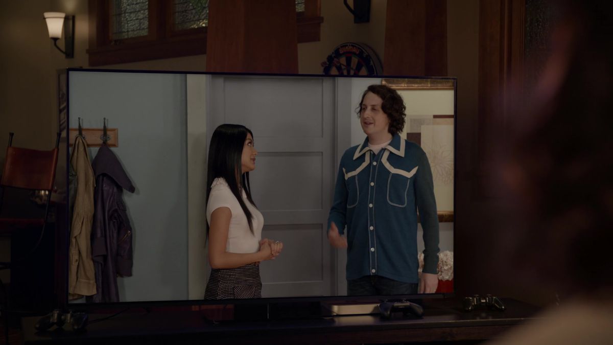 A woman with long black hair and a man with medium length, curly brown hair banter in a living room. They are depicted on a flatscreen TV.