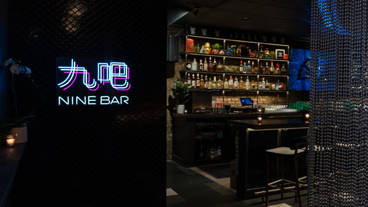 A neon sign on a black wall in front of a dark bar and a shelf full of colorful liquor bottles.