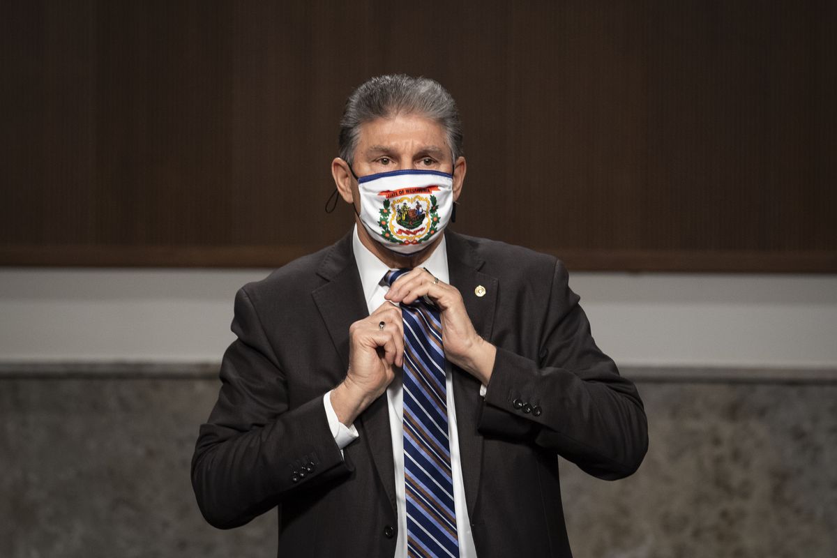 Manchin, in a dark suit, white shirt, and West Virginia flag mask, adjusts his blue and gray striped tie.