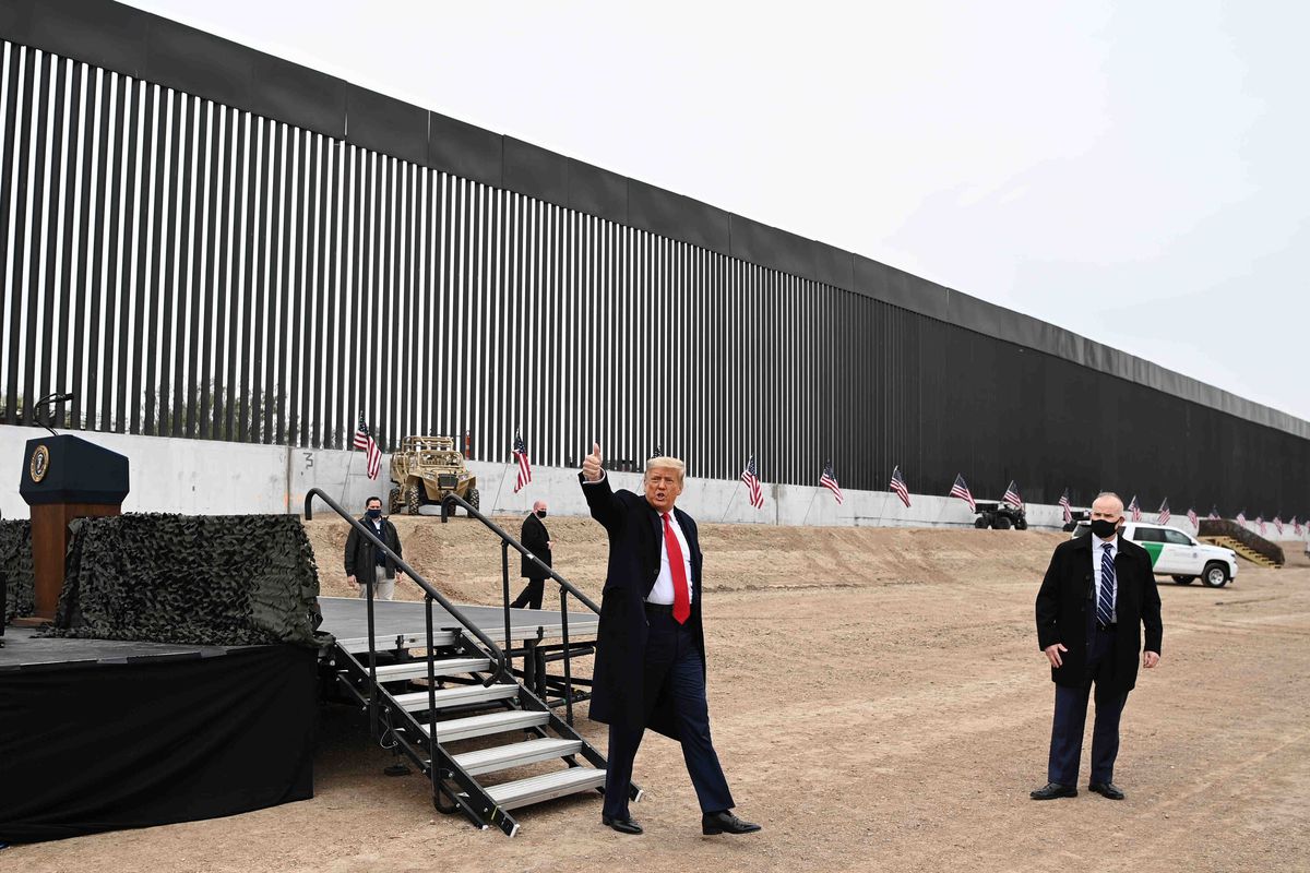 Trump, in a long unbuttoned black dress coat, wind pulling at his bright red tie, smiles and raises his right arm, giving a thumbs up. Behind him loom the massive black bars of the border wall, dark against a grey sky.