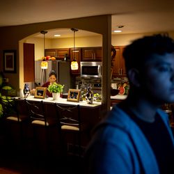 5:51 a.m.: Luis Hernandez, right, watches the news as his mother, Mariela Hernandez, cleans the kitchen before the two leave for work at an ink and toner cartridge factory. His mother and Luis work the early shift before the day’s classes so he can afford to go to college at Metropolitan State University of Denver.