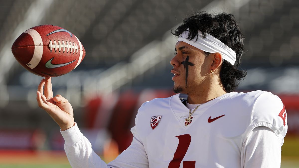 Jayden de Laura, quarterback of the Washington State Cougars, wearing a white jersey with a crimson number four, spins a football on his index finger.
