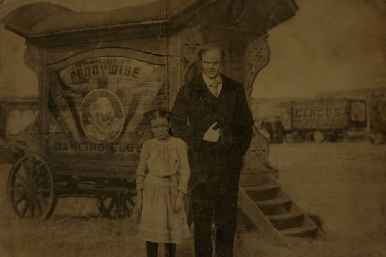 An old aged image of a girl in a white dress and a man in a black suit standing in front of a carriage painted with a sign that reads “Pennywise the Dancing Clown”