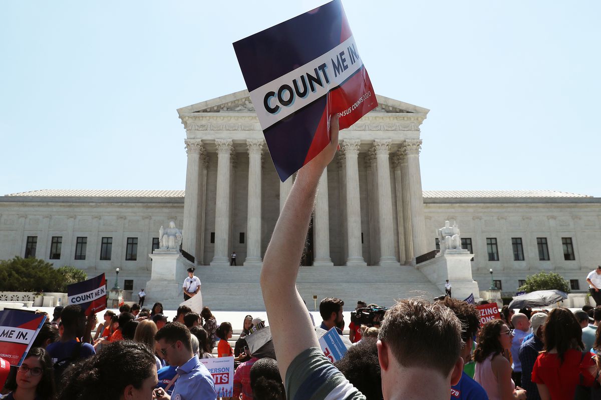 A crowd of people on the steps of the Supreme Court building, holding signs including one high in the air that reads “Count me in.”