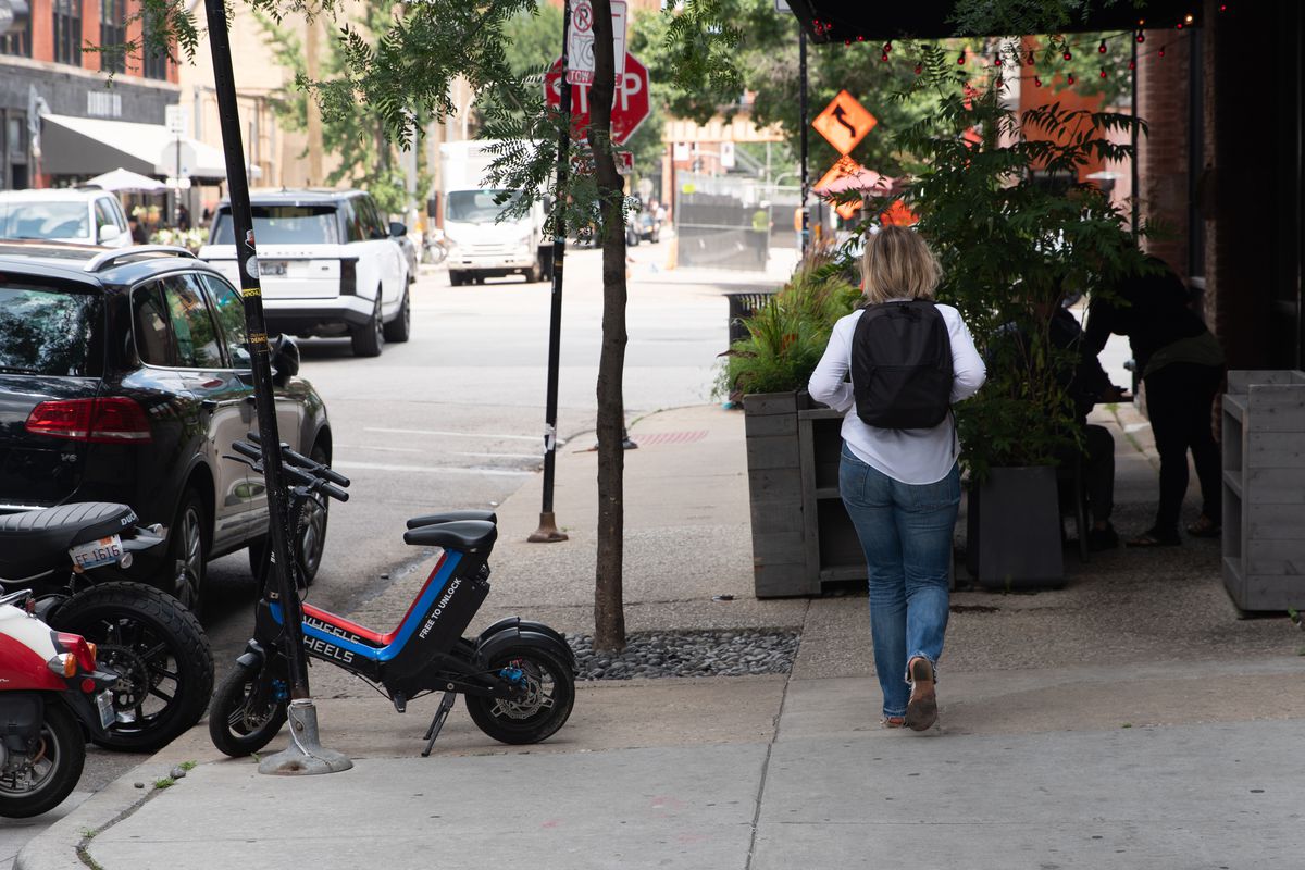 A pedestrian navigates a sidewalk crowded with cafe seating, foliage and scooters in the 100 block of North Green Street in the West Loop.