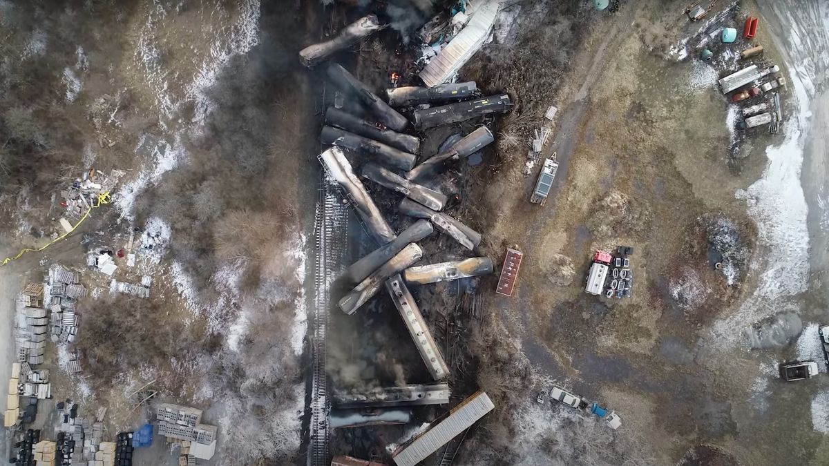 A view from above shows 20 or so train cars in disarray over the train track, most of them blackened and burned.