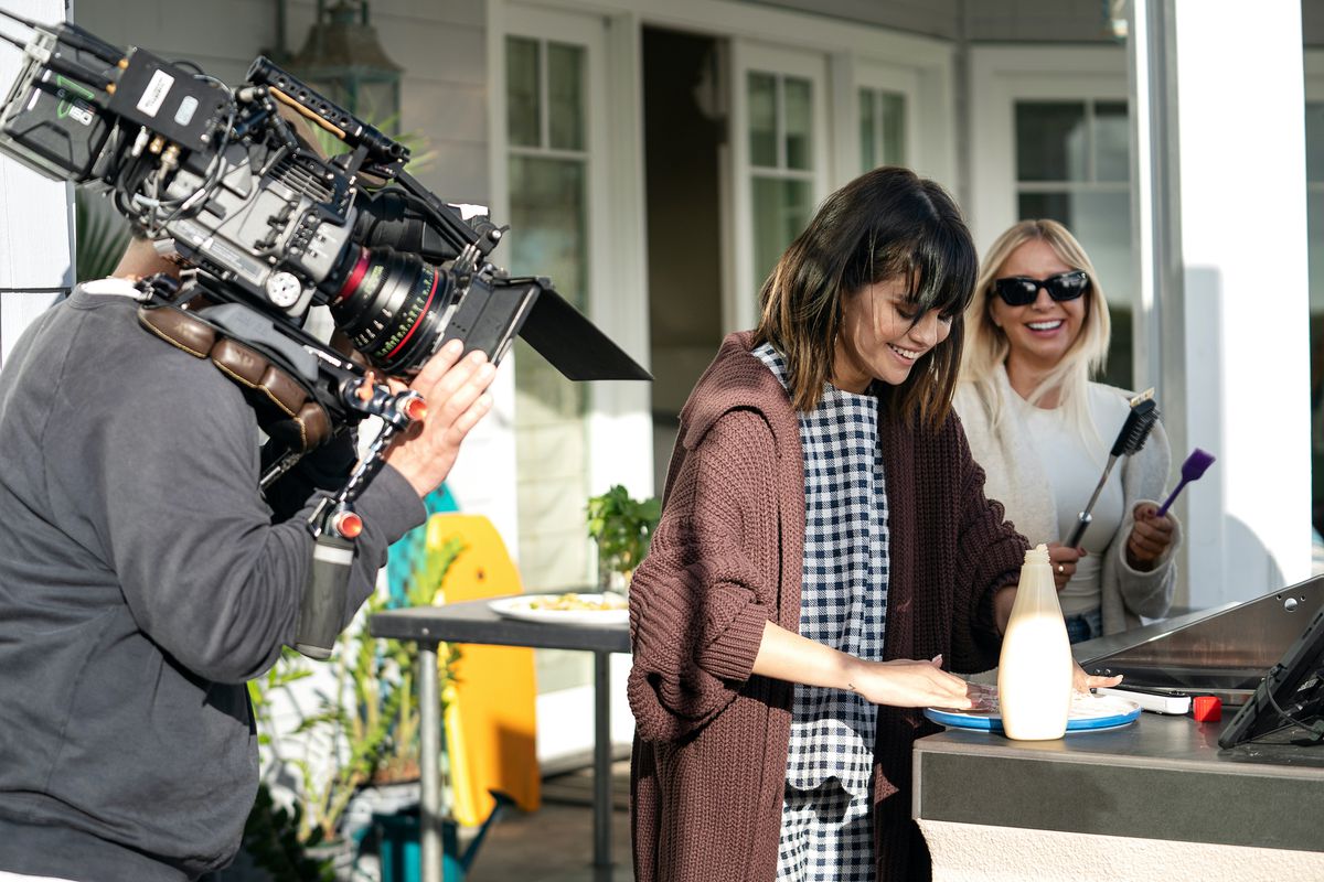 Selena Gomez, a woman with brown hair wearing a brown cardigan, stands in front of a table next to a woman wearing a hijab and sunglasses. Off to the side, a man films the scene with a camera.