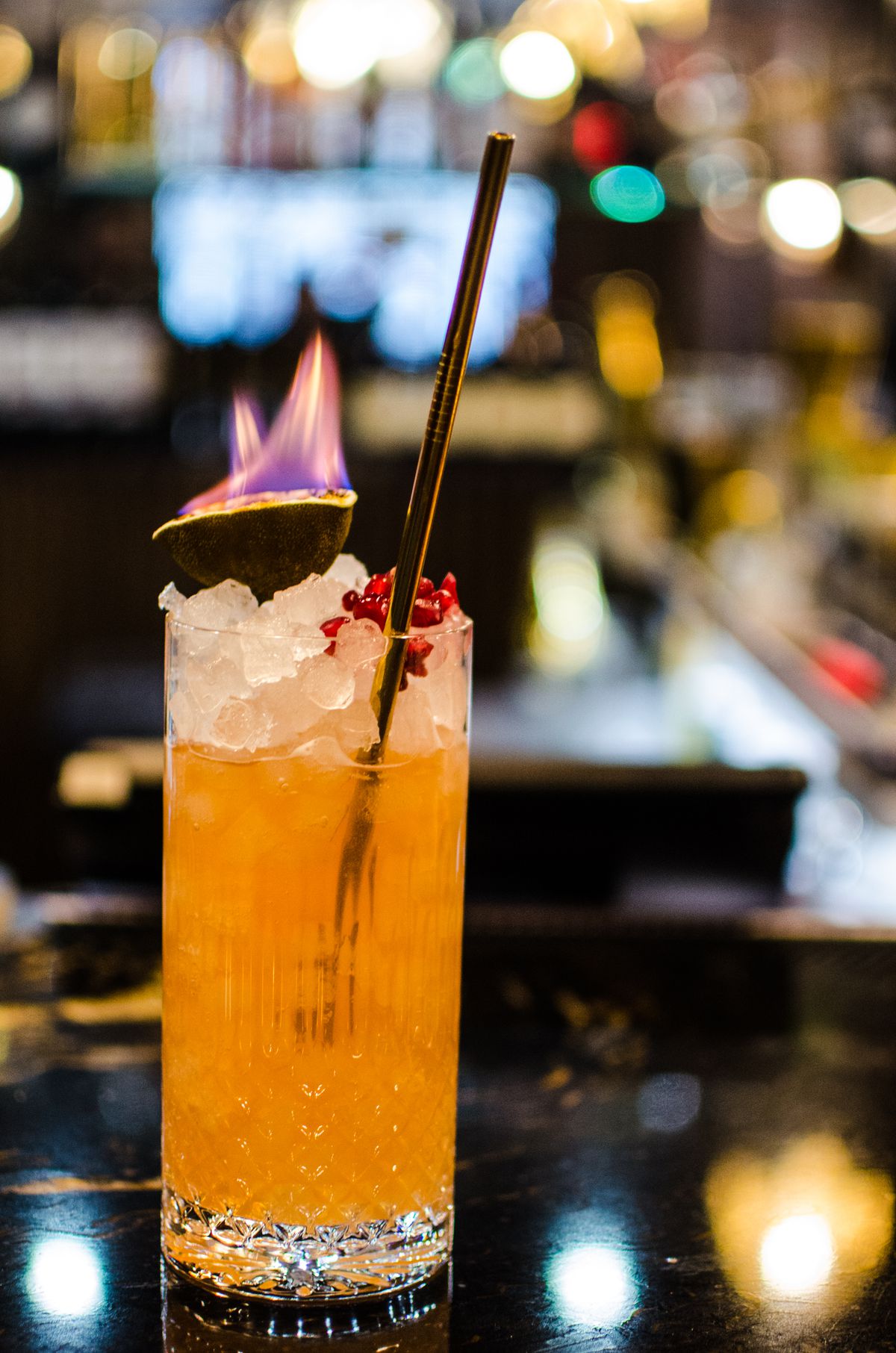 A pink-orange cocktail is topped with a lime skin on fire and pomegranate seeds.