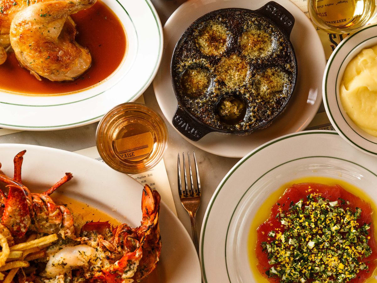 A spread of dishes sit on a table at Pastis, including golden roast chicken, buttered lobster, snails, and crimson raw tuna