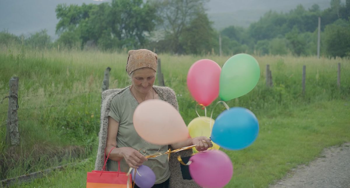 An older woman in a kerchief stands near a field, holding a bunch of balloons.