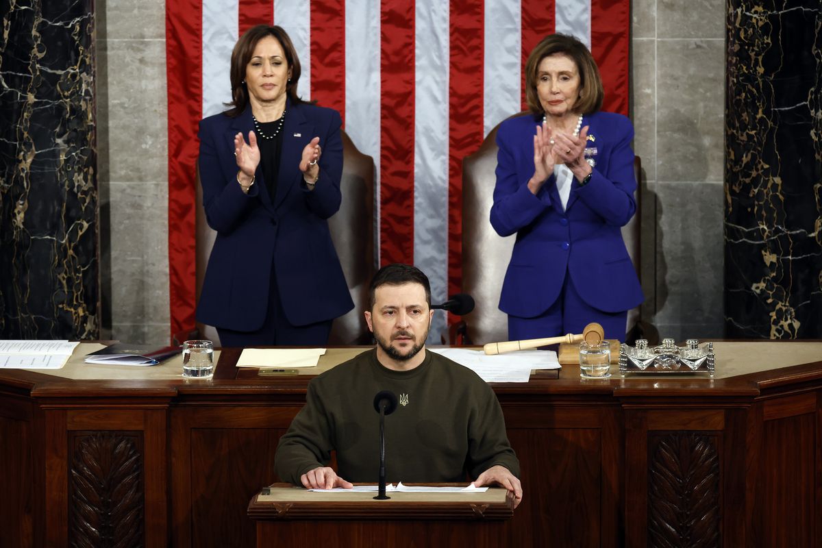 Zelenskyy, in olive fatigues, his shirt featuring the gold trident crest of Ukraine, looks serious, speaking before a podium. Behind him are Vice President Kamala Harris and House Speaker Nancy Pelosi, who are on their feet, applauding.