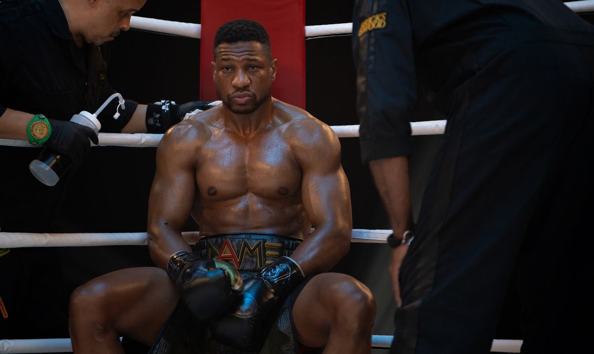 Damian (Jonathan Majors) slouches in the corner of a boxing ring during a match, shirtless and sweaty, as attendants offer him a water bottle and lean over to confer with him in Creed III