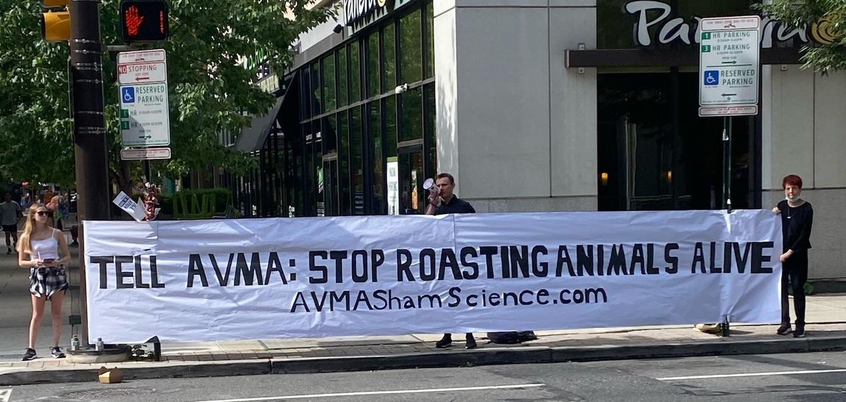 Three activists hold a large banner that reads “TELL AVMA: STOP ROASTING ANIMALS ALIVE. AVMAShamScience.com”