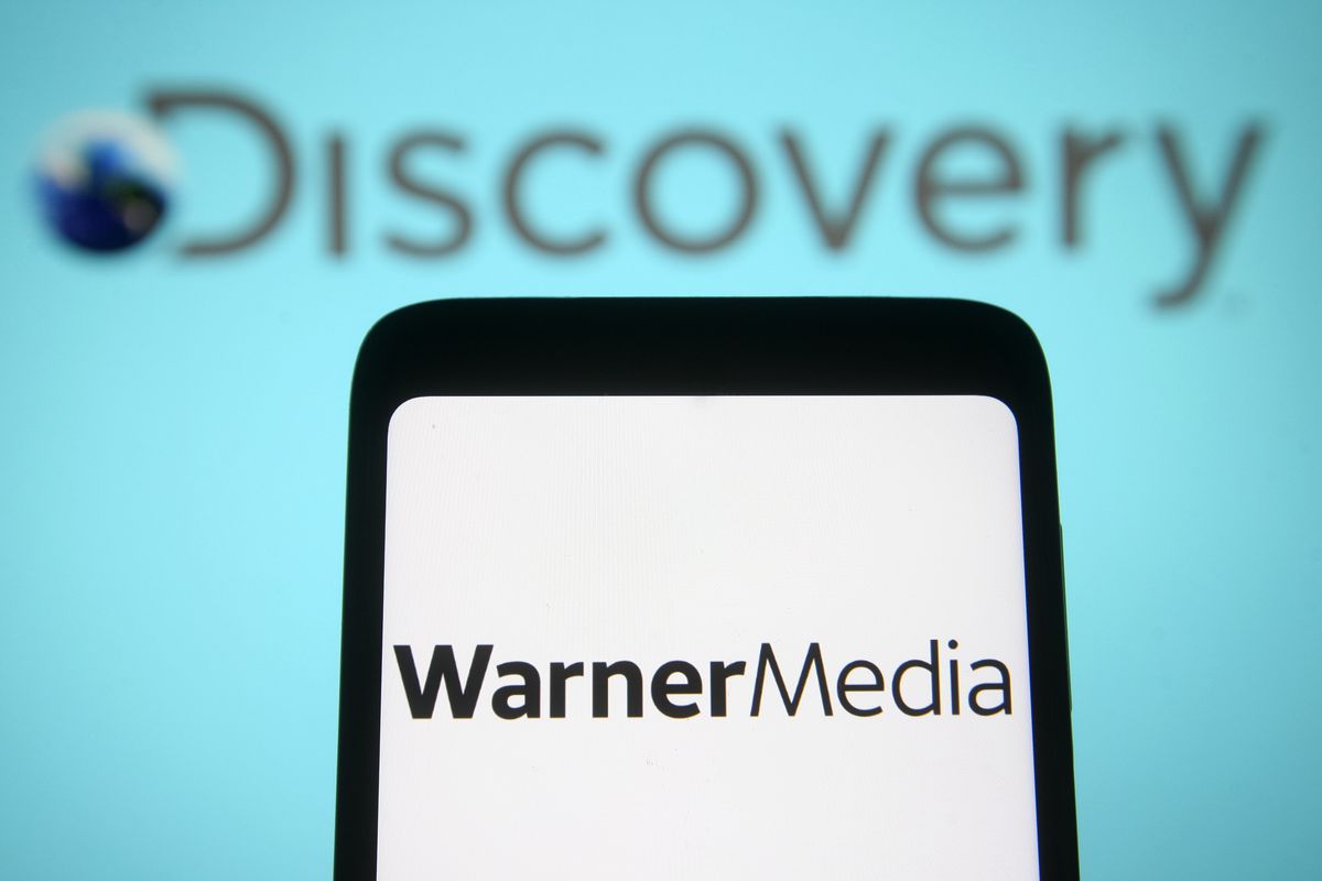 In this photo illustration, a Warner Media LLC logo is seen on a phone in front of a Discovery logo.