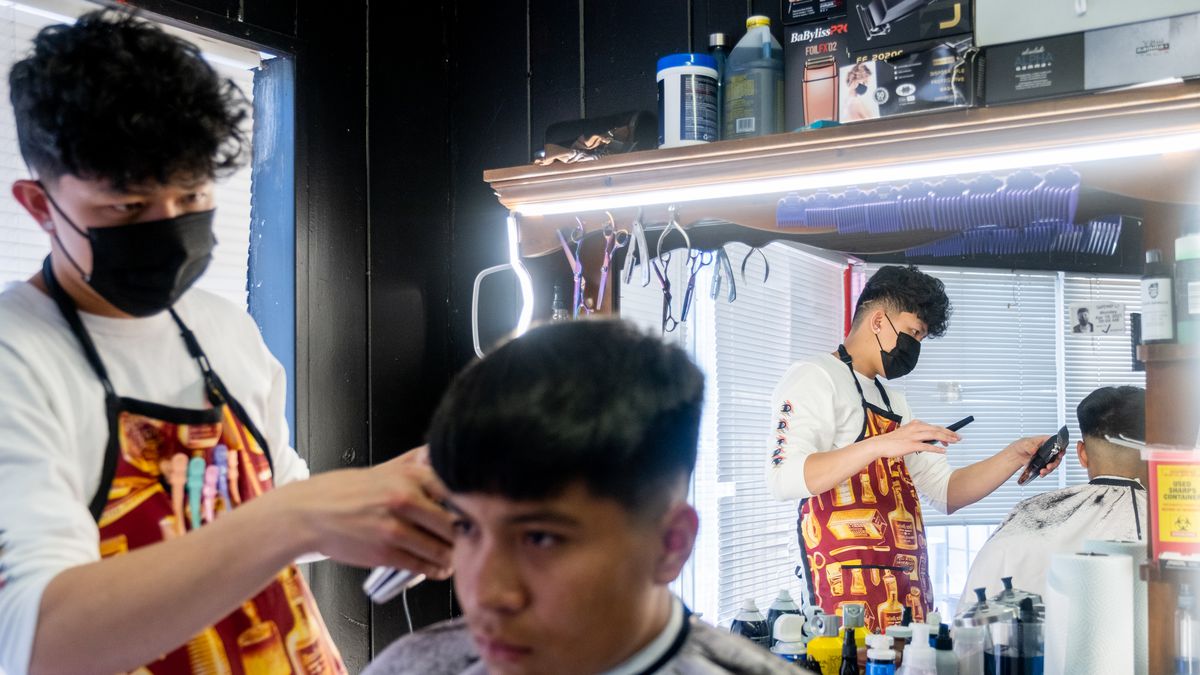 A young man wearing a black mask and barber’s apron cuts the hair of another young man in a barber shop. The barber is reflected in his mirror, adorned with bright lights at the top.