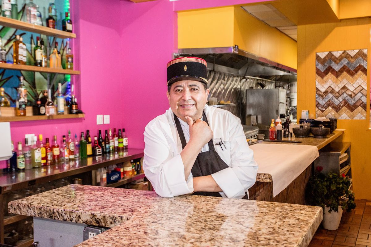 A chef in a black hat smiles and poses behind a bar.