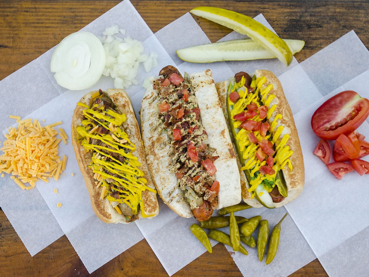 three gourmet hot dogs sit on paper on a wooden table surrounded by condiments like pickles, cheese, and peppers
