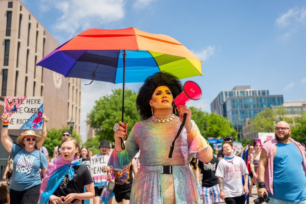 A person in a colorful dress and black wig holding up a rainbow umbrella and megaphone in front of a crowd of people holding up signs in support of the LGBTQ community.