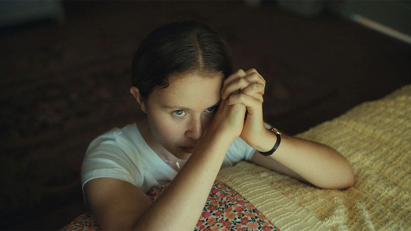 A teenaged girl has her hands folded in prayer at the edge of her bed.