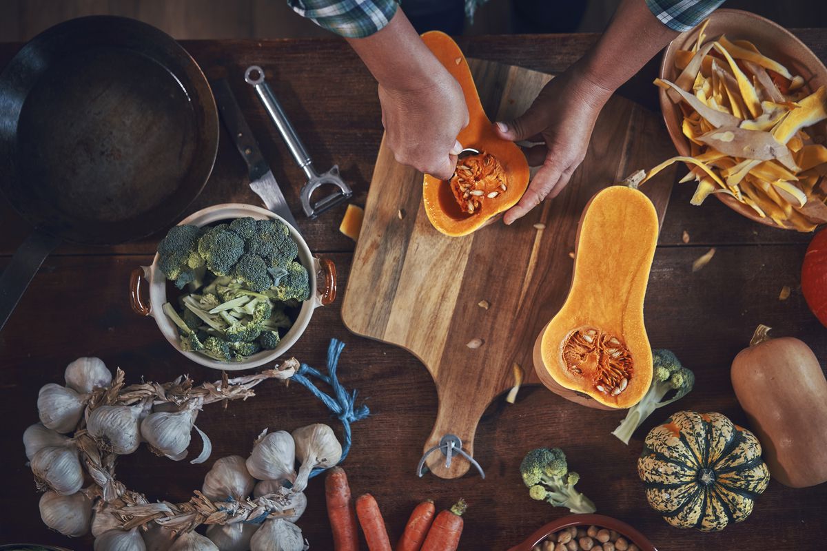 Kitchen table viewed from above strewn with garlic heads, squash, broccoli, and vegetable peelings. Hands visible preparing squash over a cutting board.