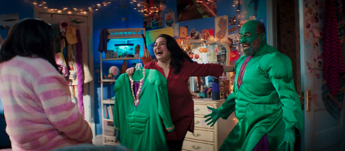 kamala khan’s mother holds up a small hulk cosplay made out of traditional pakistani clothing; her father wears a larger, matching costume,c complete with green face paint