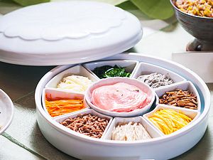 A round segmented dish with colorful, small vegetable-based dishes in each segment around a center dip with the dish’s lid set nearby