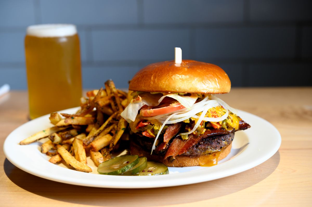 A bacon burger, fries, and sliced pickles on a white plate with a beer in the background.