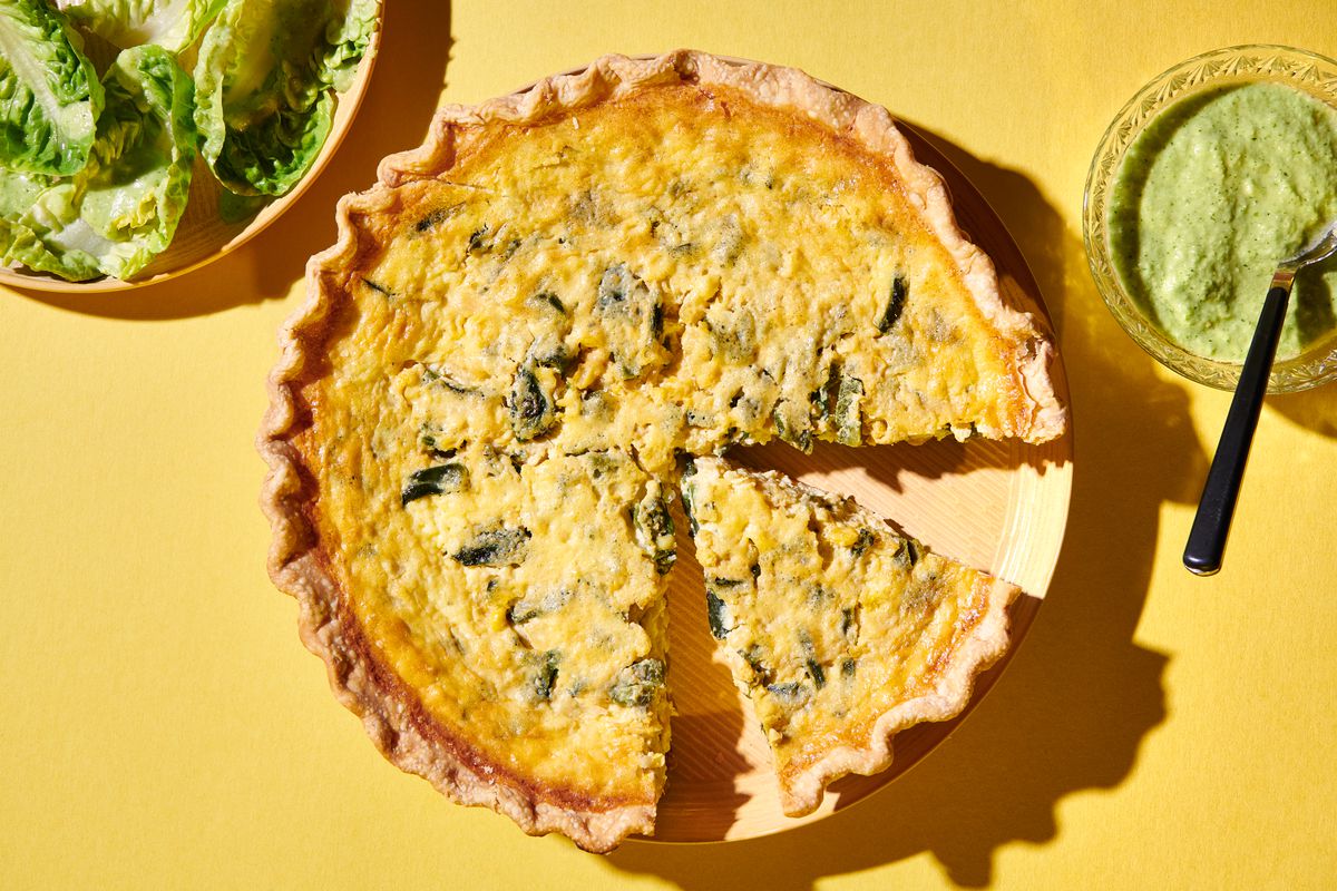 A rajas con crema quiche, served whole with a slice removed and accompanied by a plate of greens and a little bowl of green dressing.
