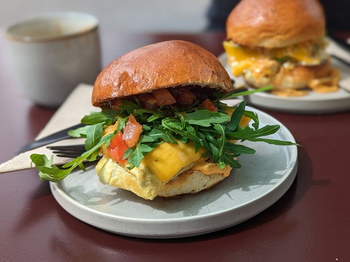 A breakfast sandwich with arugula spilling out over tomato, melted cheese, and omelet. 