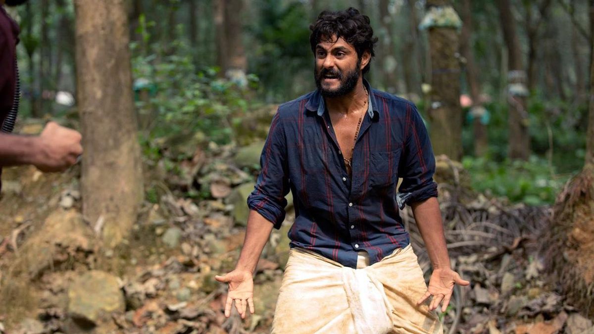 A man wearing a plaid shirt extends his hands and makes an angry face in a forest in Angamaly Diaries.