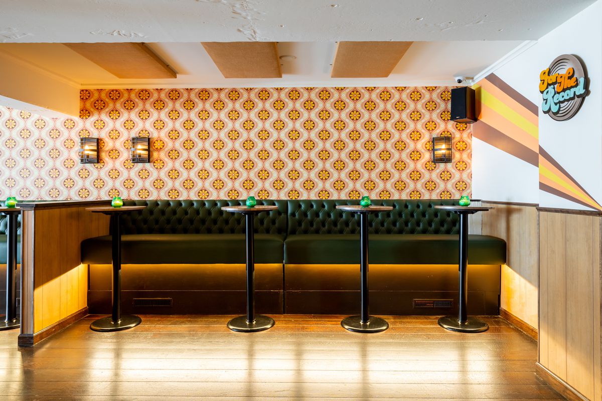 Four high-top tables in front of green banquettes against a wall with flower wallpaper.