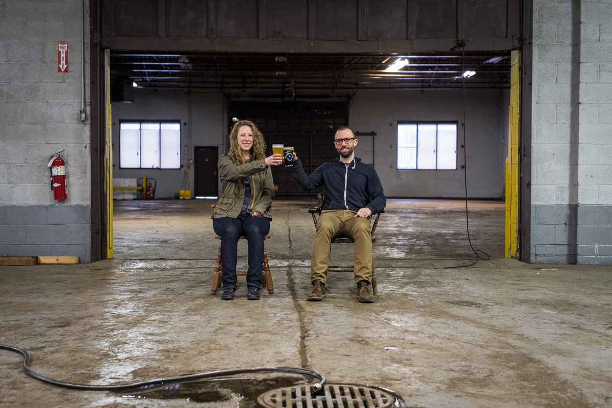 Rachel Szlaga and Paul Szlaga sit on chairs in their unfinished brewery warehouse cheersing with pints of beer. 