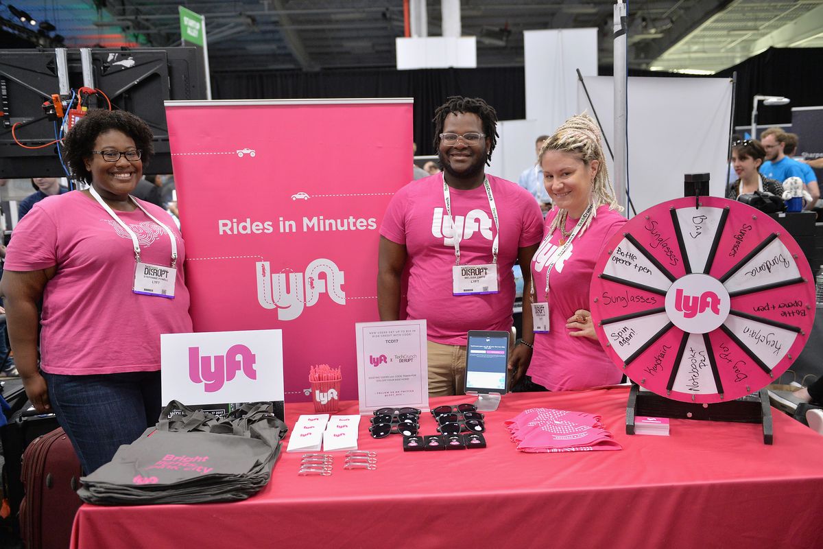Lyft employees stand at a table filled with Lyft logos and merch at TechCrunch Disrupt NY 2017. 