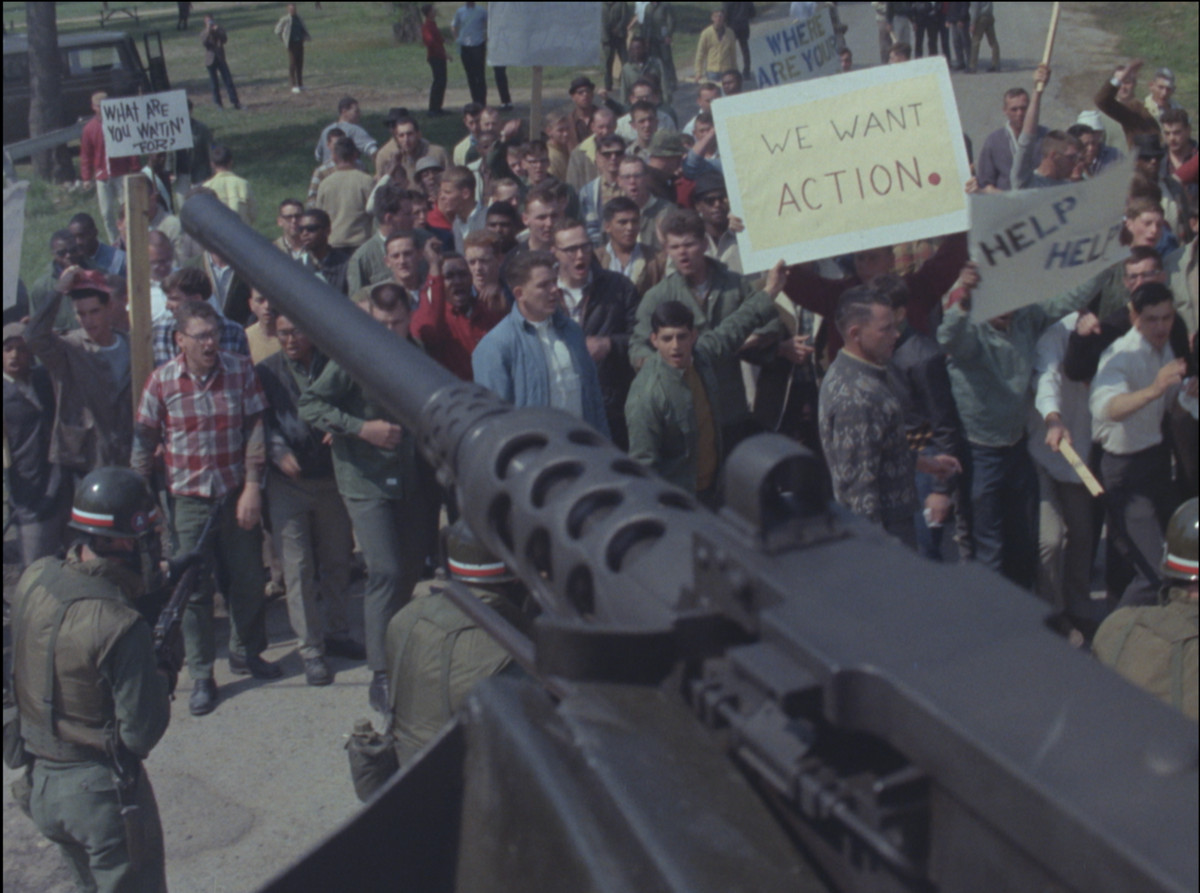 A group of protestors holding up signs while a row of soldiers stand in front of them with a turret gun aimed above the crowd.