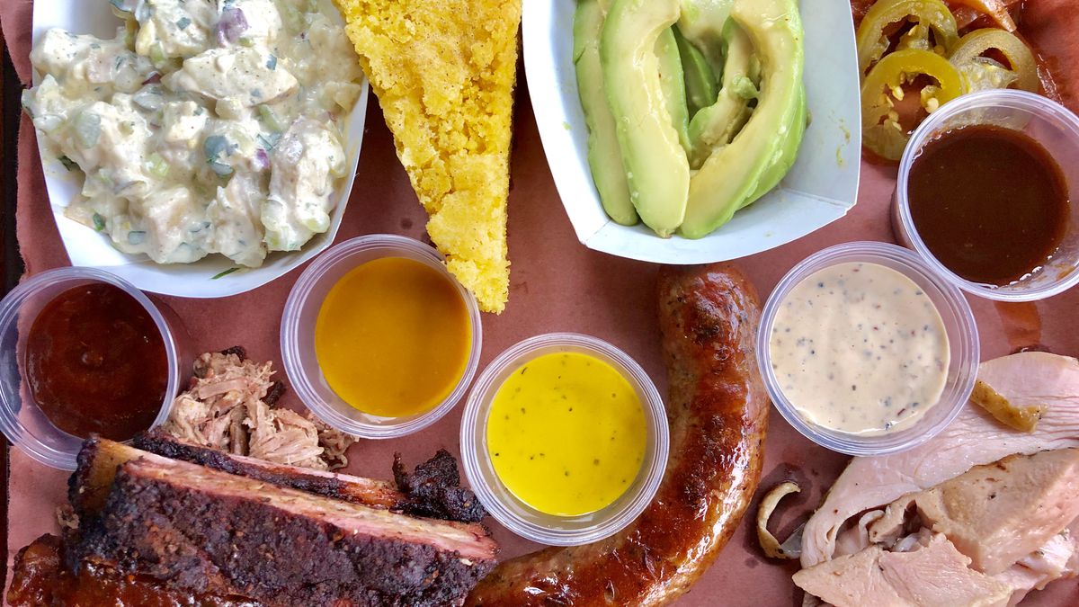 An overhead photo of a tray of barbecue, including ribs, a sausage, turkey, cornbread, pulled pork, pickled avocado, potato salad, and barbecue onions