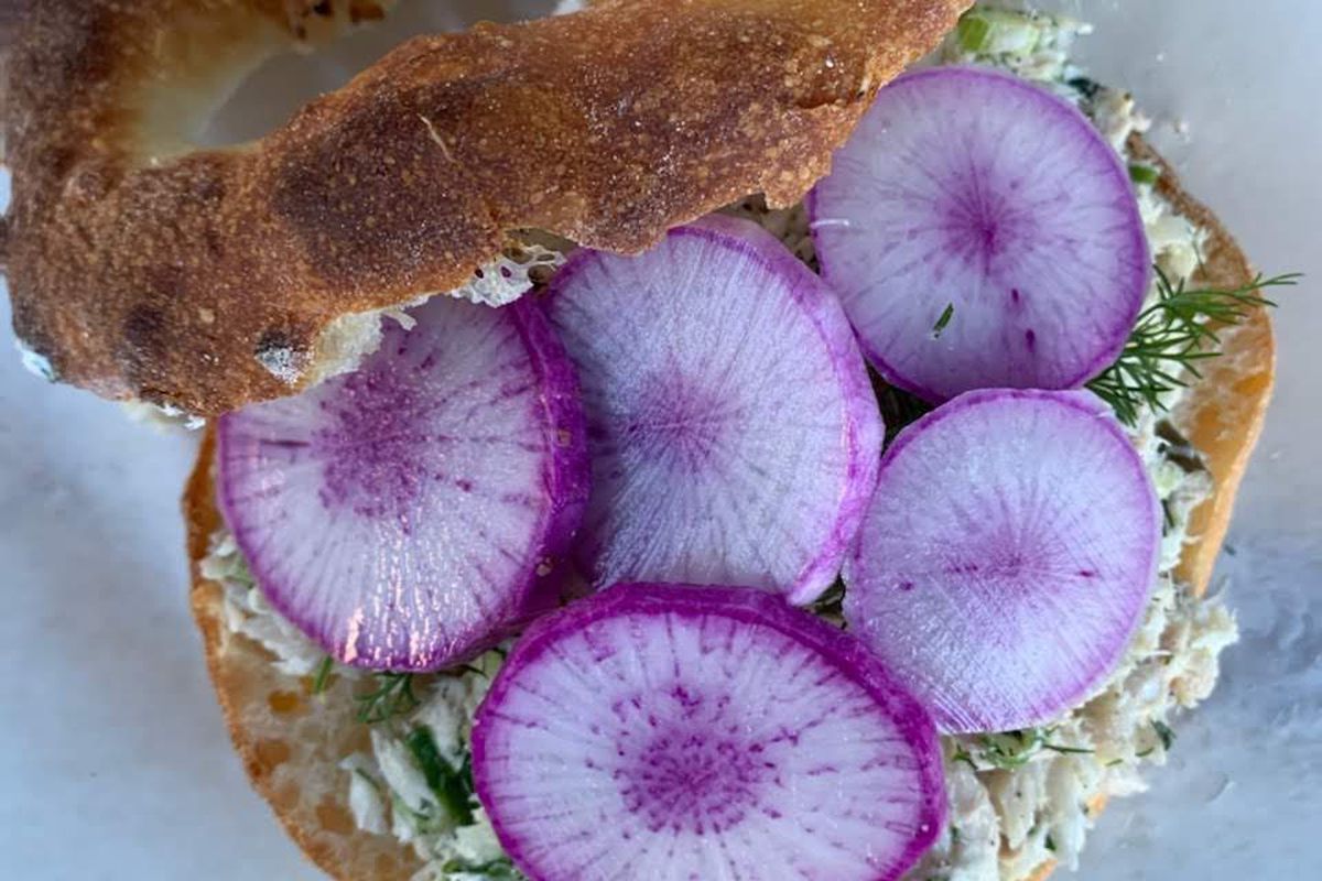 A bagel cut in half with shallot cream cheese, sprigs of dill, and thin slices of pink radishes.