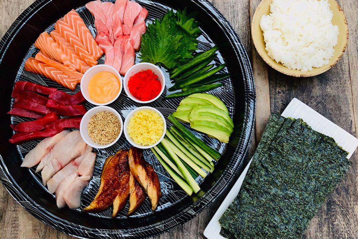 A colorful array of sliced raw fish, including salmon and tuna, along with julienned veggies on a black tray. A plate of crisped nori and bowl of rice sit to the side. 