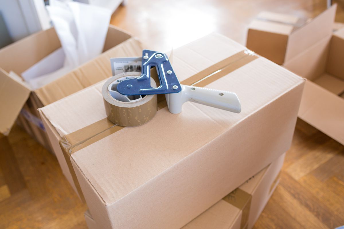 A brown cardboard moving box amongst other moving boxes inside a home with a roll of packing tape on top. 