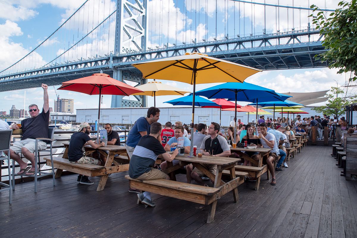 outdoor deck with people at picnic tables under colorful sunbrellas with a bridge in the background