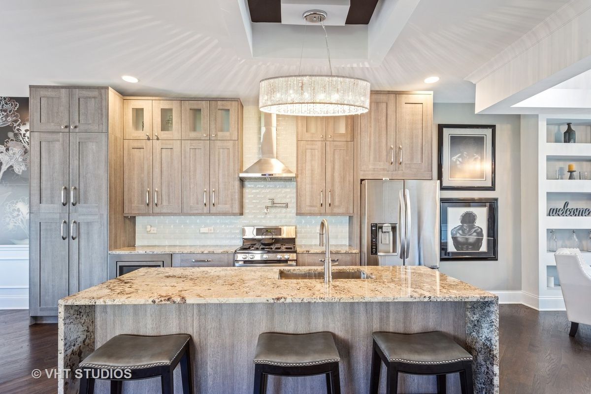 A kitchen with a stone waterfall island with three stools and gray wood cabinets and stainless appliances. 