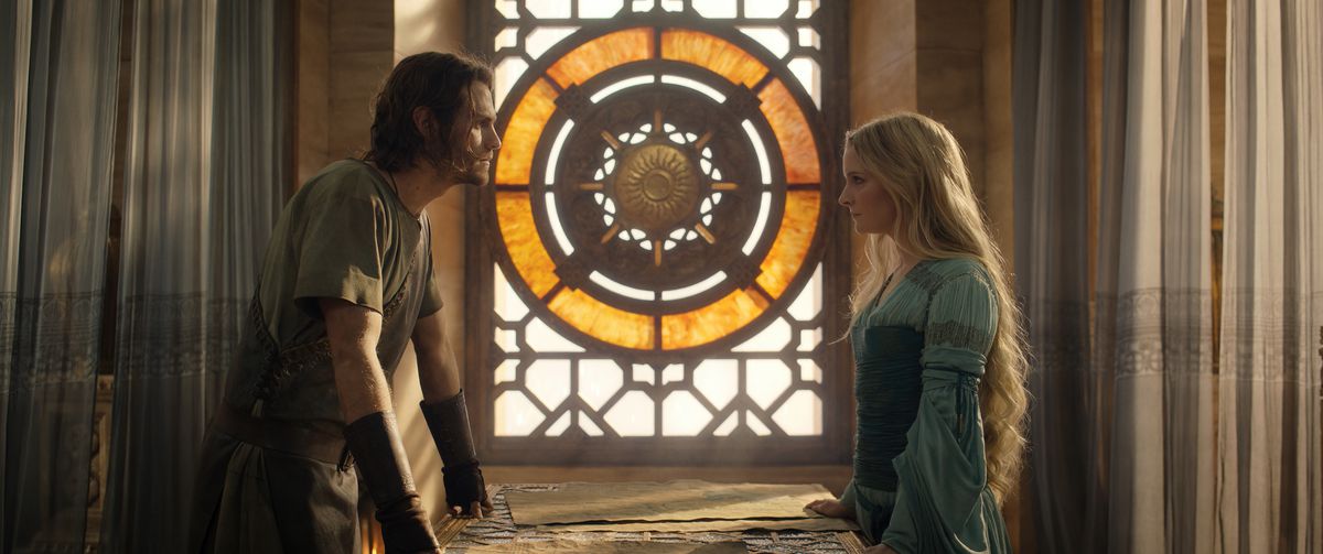 Galadriel stands across a table from Halbrand, who places his hands on it and leads toward her as an ornate window with a circular pattern illuminates the space between them both.