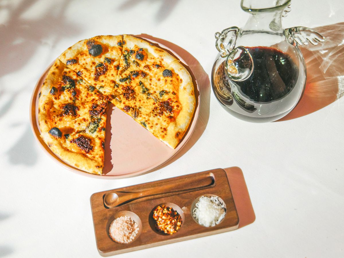 A pizza is served on a marble top with a bottle of olive oil and a tray of seasonings.