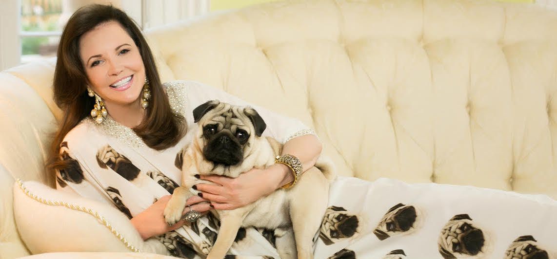 A woman reclining on a couch, holding a pug, wearing a caftan with a pug’s face on it