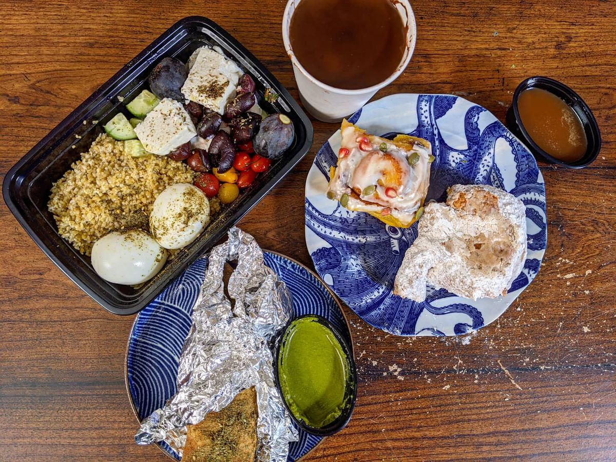 Overhead view of a wooden table full of breakfast takeout items: two pastries sit on a blue-and-white octopus plate, a paper cup holds a thick hot chocolate, and more