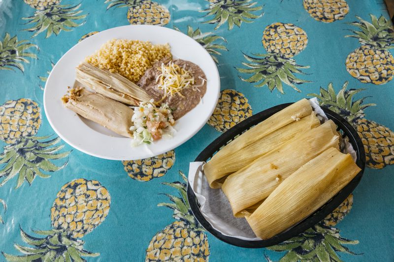 One white plate with a tamale alongside rice and beans sits next to a plastic basket of three unwrapped tamales.