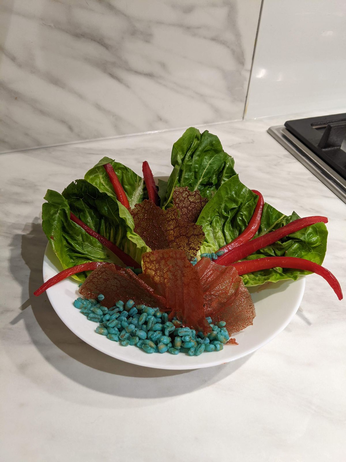 A salad on a plate with leafy greens, red-dyed carrots, and a small pile of blue grain.