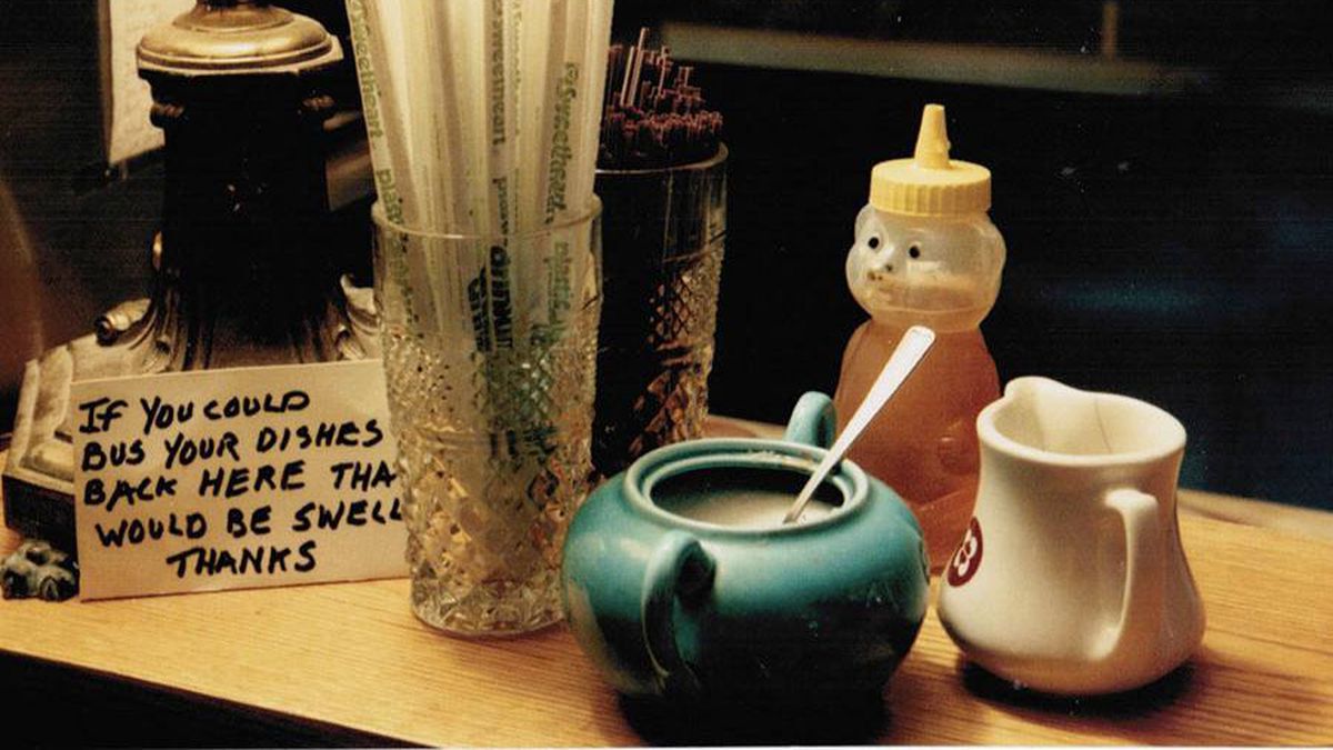 Sugar, cream, a bear-shaped squeeze bottle of honey, and straws sit on a cafe counter next to a sign that reads, “If you could bus your dishes back here that would be swell thanks.”